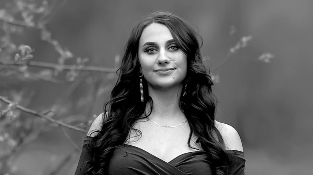 Weronika zmarła nagle Młoda kobieta o długich, ciemnych włosach, ubrana w elegancką suknię z odkrytymi ramionami, stoi na tle rozmytego naturalnego krajobrazu. Kobieta delikatnie się uśmiecha, ma na sobie długie kolczyki i subtelny naszyjnik.