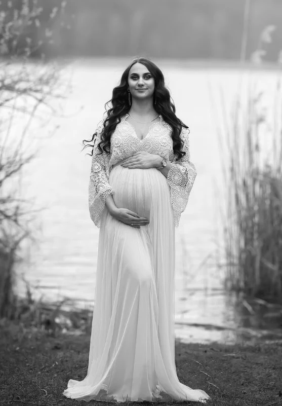 Kobieta w ciąży ubrana w długą, zwiewną suknię stoi nad brzegiem jeziora, trzyma ręce na zaokrąglonym brzuchu, w tle roślinność i delikatnie zarysowana linia wody.