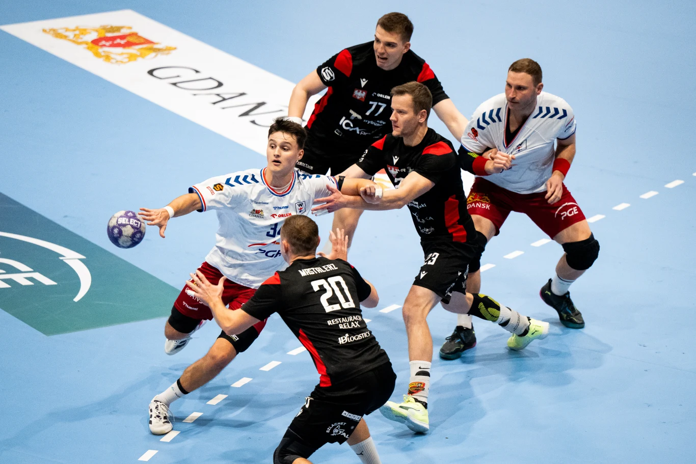 Zawodnicy dwóch drużyn piłki ręcznej walczą o piłkę na boisku, wszyscy skoncentrowani na akcji, dynamiczne ustawienie zawodników, w tle fragment niebieskiego parkietu oraz logo klubu.