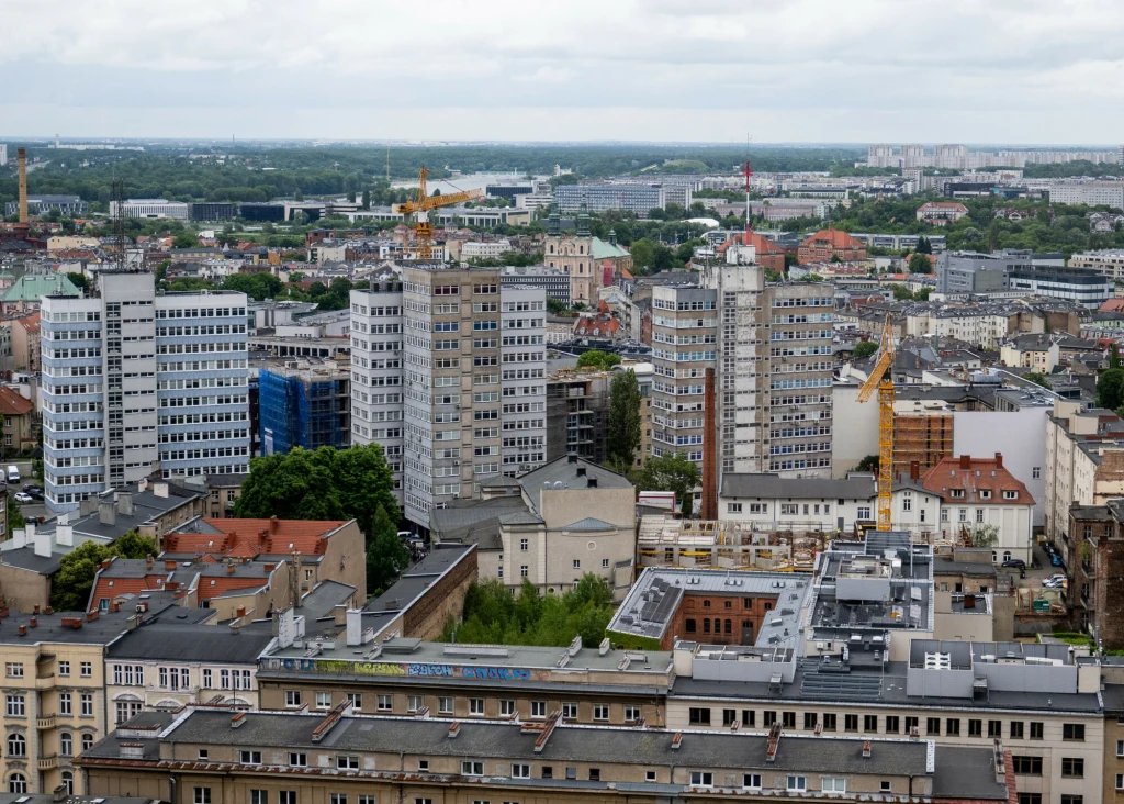 Panorama miasta z widocznymi nowoczesnymi oraz starszymi budynkami, kilka żurawi budowlanych sugeruje trwające inwestycje, wielu oknach odbija się światło, w tle zieleń i rozległe przedmieścia.