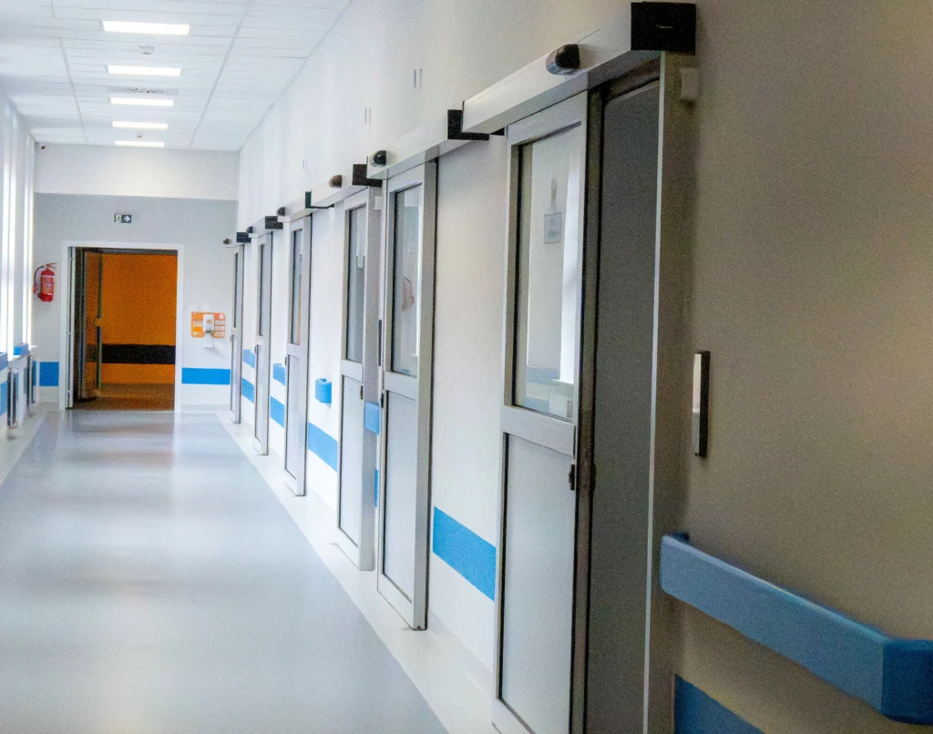 Długi, jasny szpitalny korytarz z szeregiem zamkniętych drzwi do sal, błękitnymi poręczami po bokach ścian oraz białym, czystym sufitem z oświetleniem sufitowym.