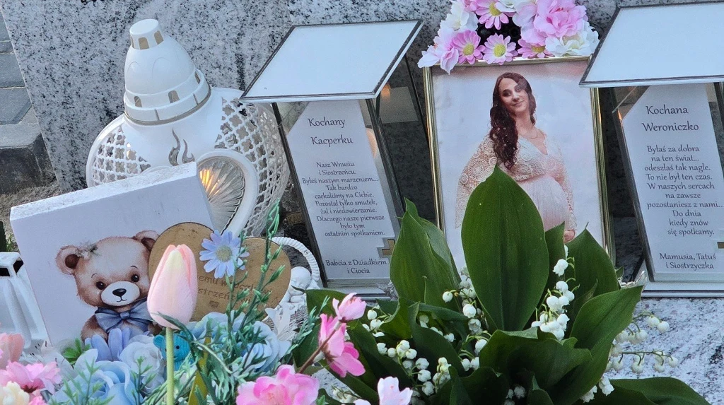 Grób ozdobiony kwiatami, zniczem i zdjęciem kobiety w ciąży, pośród dekoracji znajdują się dwa wiersze oraz obrazek misia i różowe kwiaty.