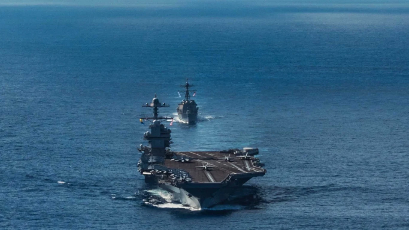 Na pierwszym planie lotniskowiec płynący po spokojnej, błękitnej wodzie, za nim podąża okręt wojenny, oba statki poruszają się po oceanie pod błękitnym niebem.