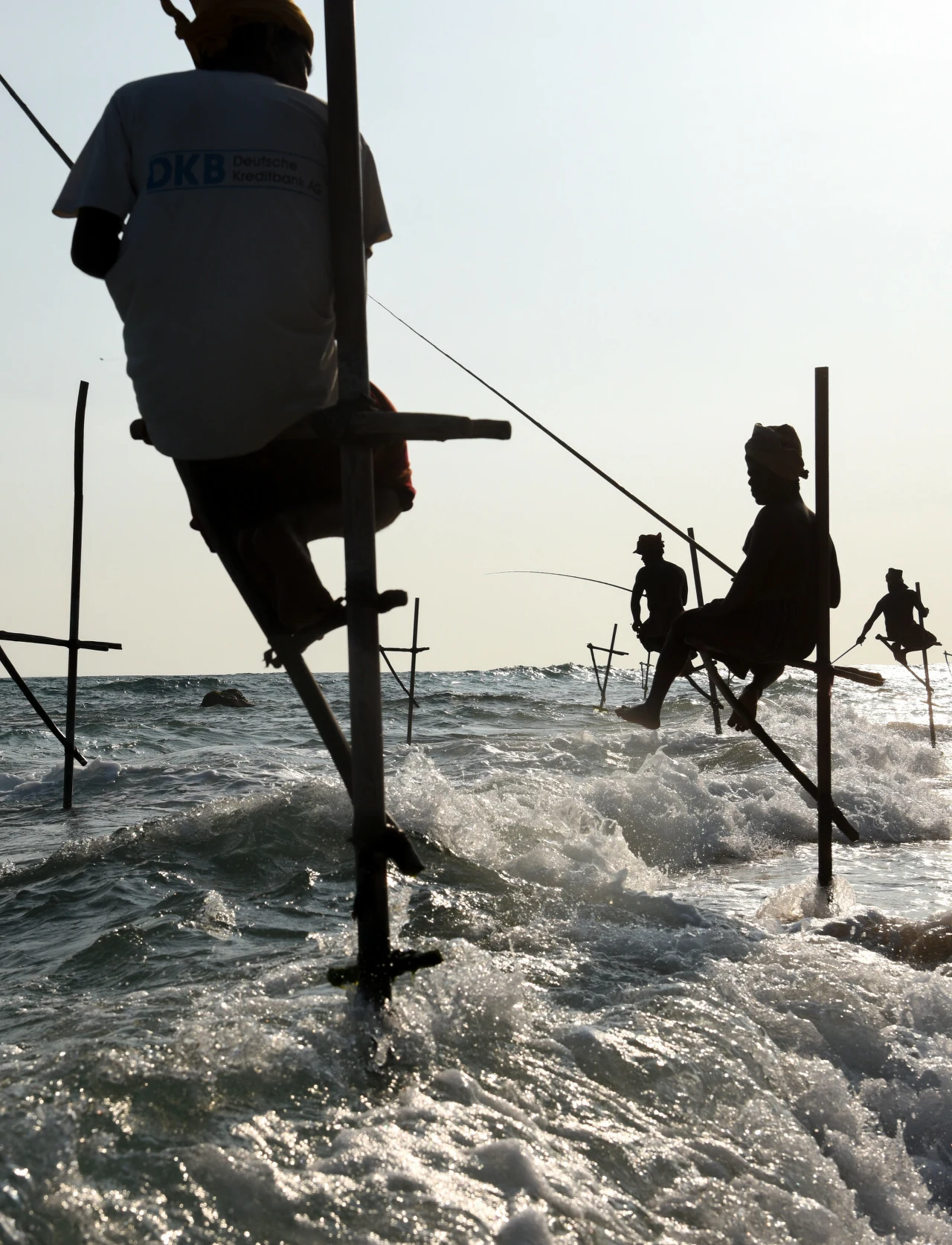 Rybacy z Cejlonu łowią ze specjalnych tyczek Rybacy siedzący na wysokich palach ustawionych w wodzie, trzymają w rękach wędki i łowią ryby, wokół nich rozbija się morska fala, sylwetki wyraźnie odcinają się na tle jasnego nieba.