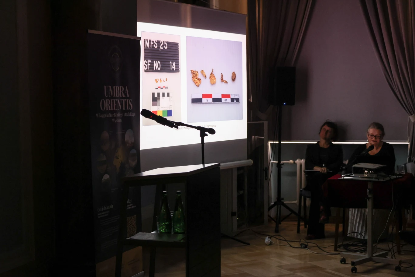 Na spotkaniu w Warszawie zespół polskich archeologów omówił badania mastaby i wyjaśnił szczegóły przygotowań do kolejnej ekspedycji. Sala konferencyjna z ustawionym ekranem, na którym prezentowane są zdjęcia archeologicznych lub naukowych znalezisk, obok ekranu ustawione są mikrofon, stolik z wodą oraz dwie siedzące kobiety przygotowane do prelekcji.