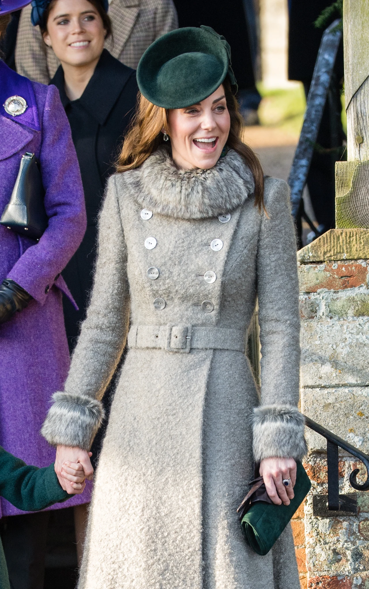 Elegancko ubrana kobieta w długim, szarym płaszczu z futrzanymi wykończeniami, zielonym beretem i zamszową torebką, uśmiechnięta, trzymająca za rękę dziecko; w tle inne osoby, w tym kobieta w fioletowym płaszczu.