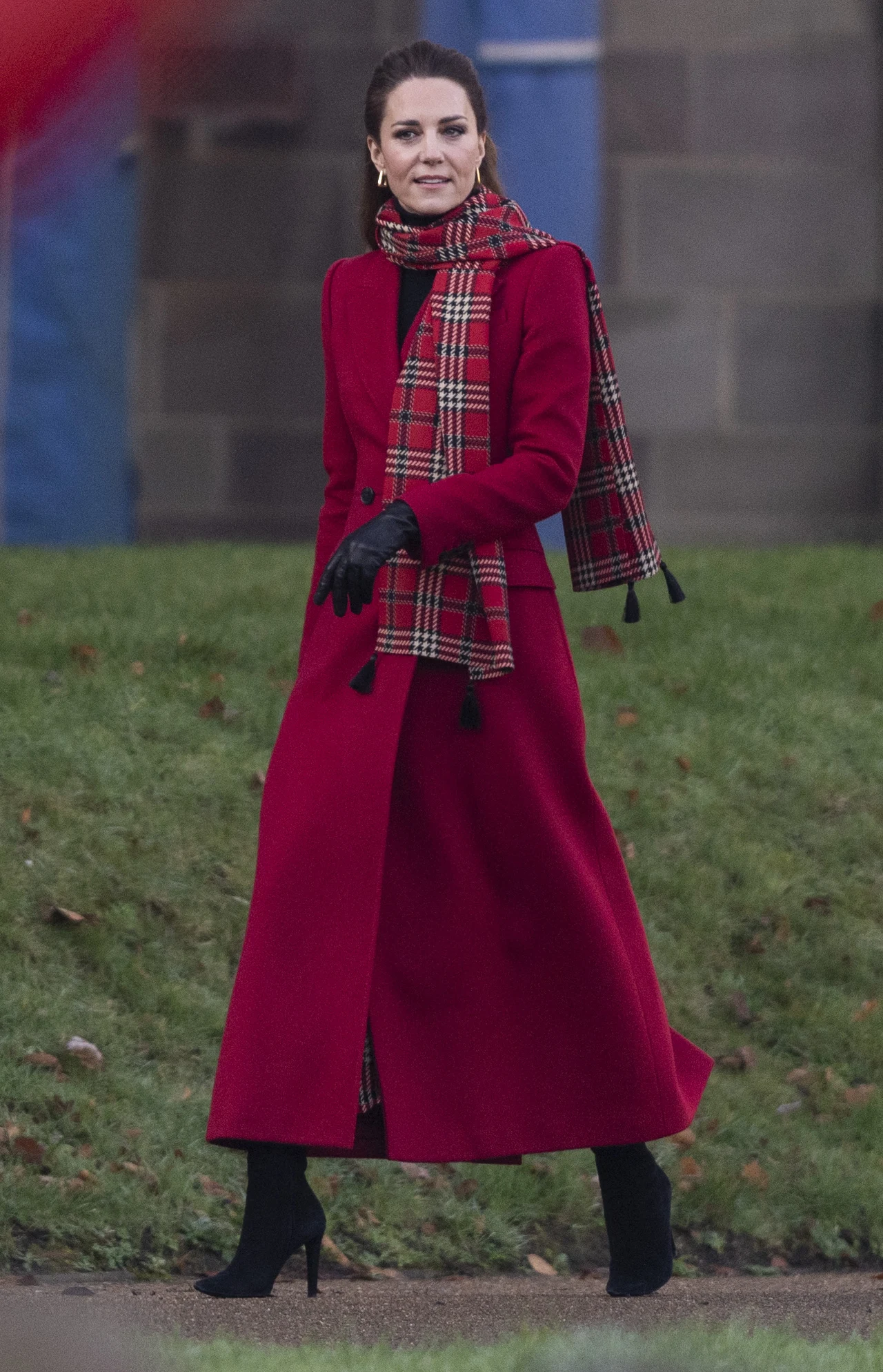 Kobieta ubrana w długi, czerwony płaszcz i szalik w szkocką kratę, stojąca na trawie przy kamiennym budynku, ma na sobie czarne rękawiczki i buty na obcasie.