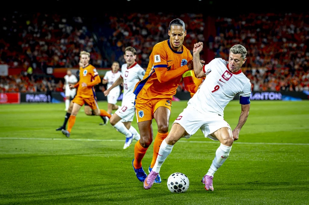 Holendrzy mrożą szampany przed meczem z Polską. Cel jest jasny, mówią o rewanżu Zawodnik w białym stroju próbuje opanować piłkę na tle dynamicznej rywalizacji z piłkarzem w pomarańczowym stroju, podczas meczu piłkarskiego na stadionie wypełnionym kibicami.