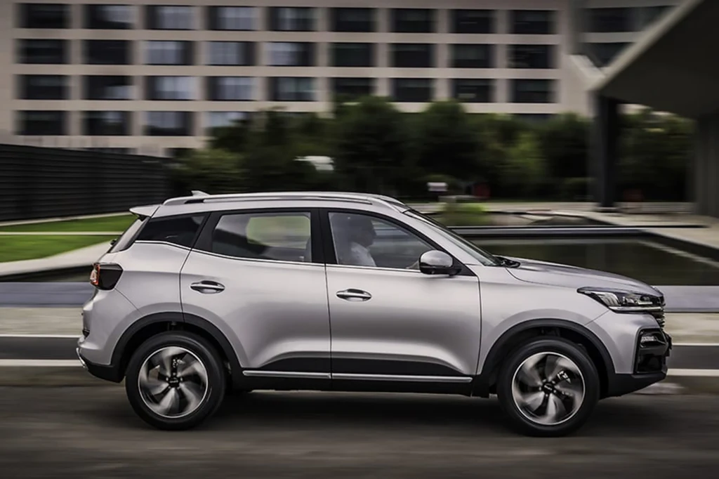 EVO Cuatro to odpowiednik Chery Tiggo 4. Jest jednak dużo tańszy Nowoczesny, srebrny SUV jadący po miejskiej drodze, w tle widoczne szare budynki biurowe i niewielki zbiornik wodny, samochód znajduje się w centralnej części kadru, podkreślając swój dynamiczny charakter.