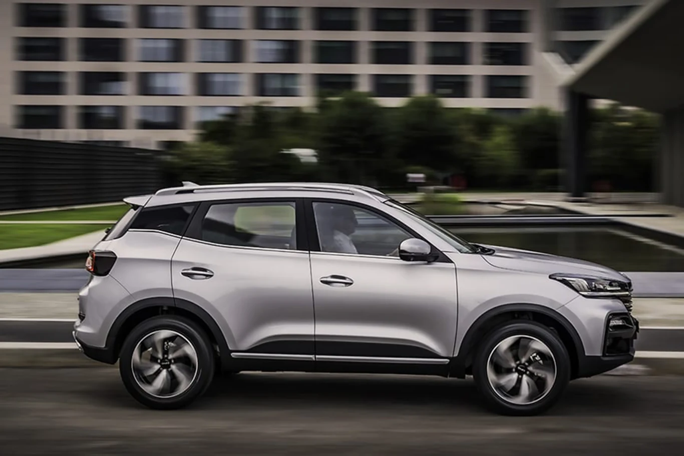 Nowoczesny, srebrny SUV jadący po miejskiej drodze, w tle widoczne szare budynki biurowe i niewielki zbiornik wodny, samochód znajduje się w centralnej części kadru, podkreślając swój dynamiczny charakter.