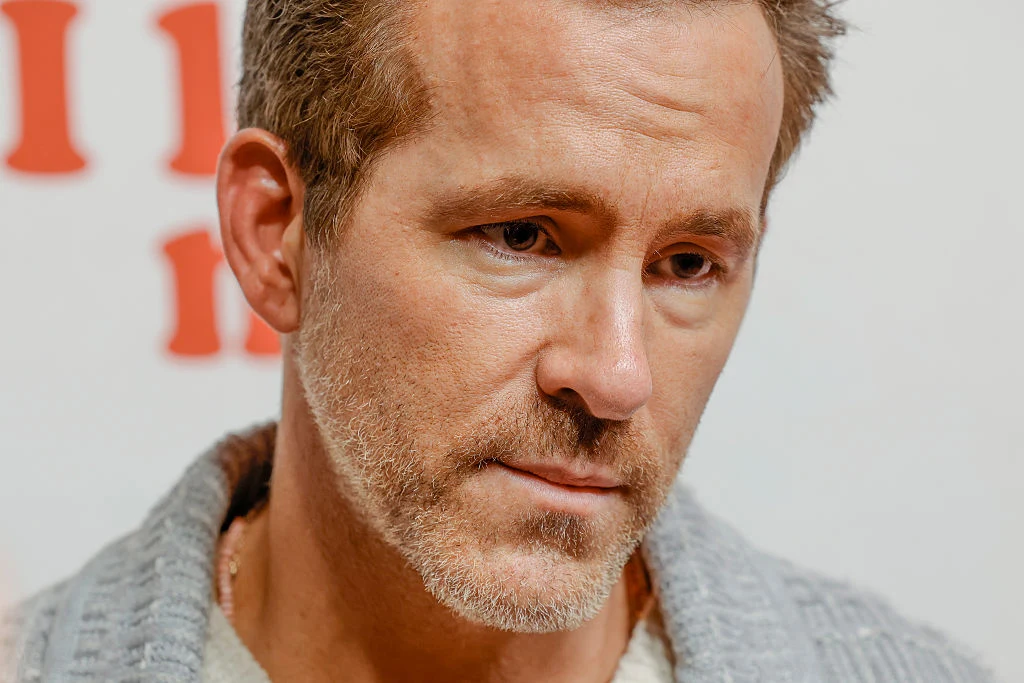 Ryan Reynolds Portret mężczyzny o krótkich, jasnobrązowych włosach i brodzie, ubrany w szary sweter, patrzący poważnie w dół na tle jasnej ściany z czerwonymi literami.