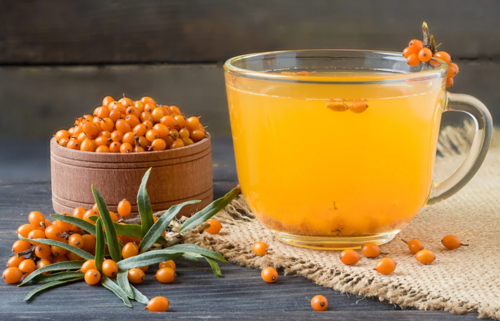 Suszone owoce rokitnika świetnie uzupełniają smak herbaty Szklanka żółtopomarańczowego soku z rokitnika ustawiona na jutowym materiale obok świeżych owoców rokitnika wysypanych w drewnianym pojemniku. W tle widoczny jest ciemny stół oraz liście rokitnika.