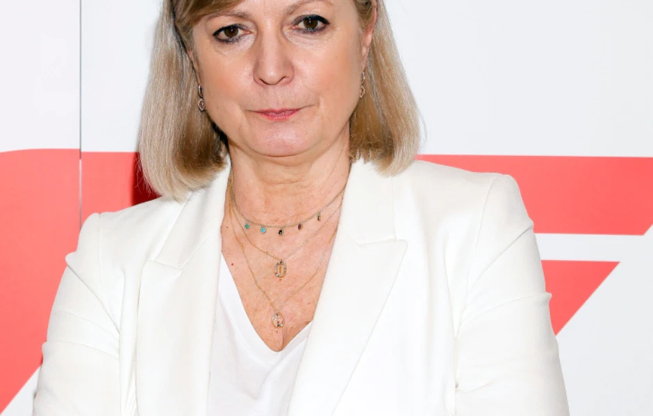 Kobieta o jasnych włosach w średnim wieku ubrana w elegancki biały żakiet i białą koszulkę, nosząca złote naszyjniki, stoi na tle białej ściany z czerwonym geometrycznym motywem.