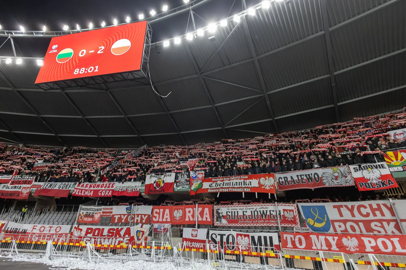 Kibice na stadionie otoczeni czerwono-białymi flagami i transparentami, wspierający polską reprezentację piłkarską. Na wyświetlaczu widoczny wynik meczu Węgry 0 Polska 2 oraz czas meczu 88:01. Stadion zapełniony kibicami, atmosfera pełna emocji sportow...