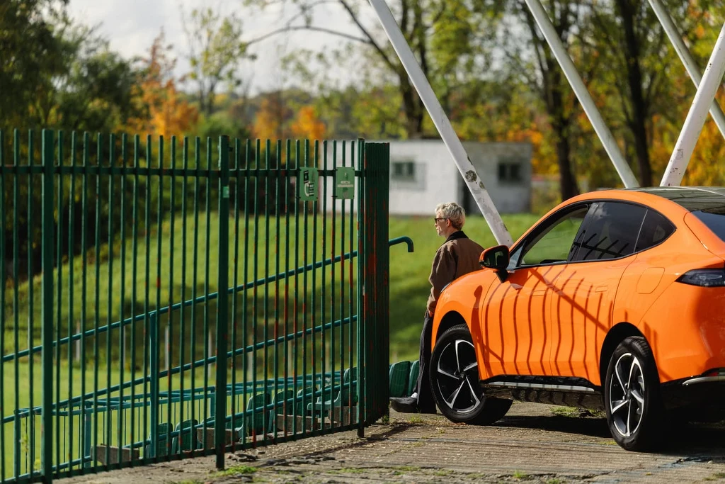 Pomarańczowy samochód typu SUV zaparkowany przy metalowym ogrodzeniu, obok którego stoi starsza osoba w okularach. W tle widoczne są drzewa z jesiennymi liśćmi oraz fragmenty budynków.
