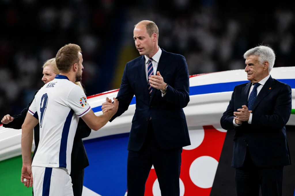 Harry Kane i książę William Zawodnik w białej koszulce piłkarskiej ściska dłoń elegancko ubranego mężczyzny, obok stoją inni oficjele w garniturach, a w tle znajduje się wielobarwny element dekoracyjny związany z wydarzeniem sportowym.