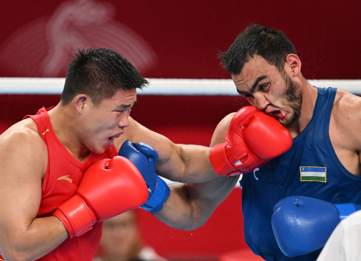 Lazizbek Mullojonov (po prawej stronie) Dwóch bokserów podczas intensywnej wymiany ciosów na ringu. Jeden z zawodników w niebieskim stroju otrzymuje silny cios od przeciwnika w czerwonym stroju. W tle widoczny fragment ringu i rozmazane tło.