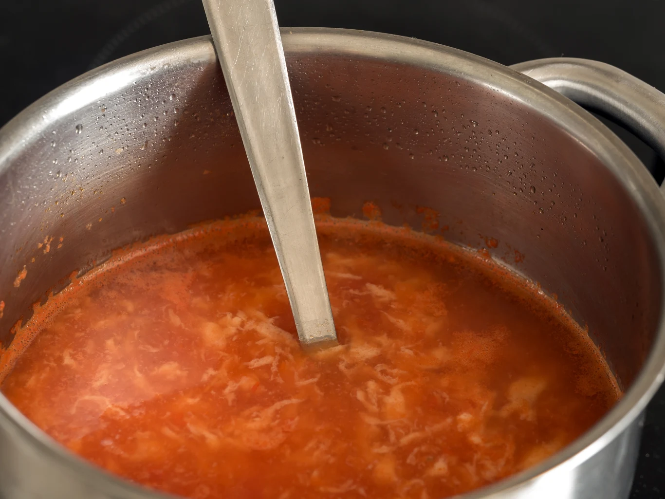 Garnek wypełniony gotującą się zupą pomidorową, w której znajduje się metalowa chochla. Na powierzchni zupy widać delikatne ślady śmietany lub jajka.