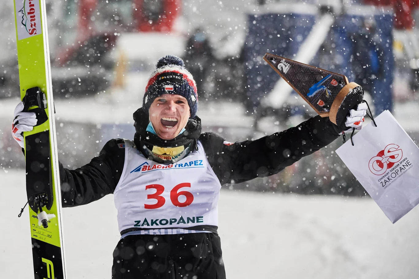 Andrzej Stękała Sportowiec w białym stroju z numerem 36, w czapce i rękawiczkach, trzyma w jednej ręce narty, a w drugiej puchar oraz torbę z logo sponsora, dookoła pada śnieg, w tle rozmyte sylwetki i sprzęt.