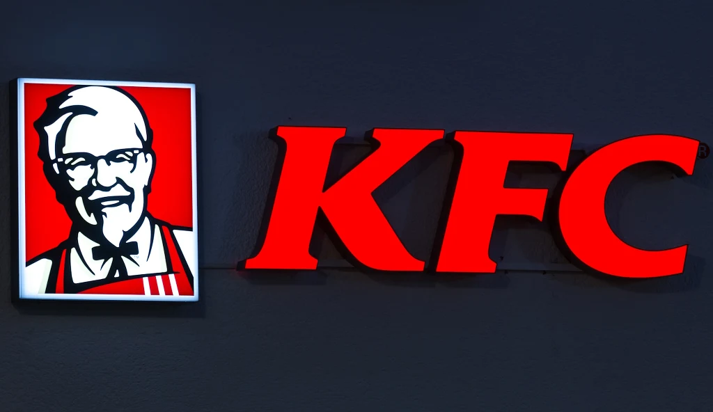 Nowa promocja w KFC to ukłon w stronę fanów popularnej gry Monster Hunter Wilds Podświetlone logo marki KFC na ciemnym tle, obok wizerunku charakterystycznego pułkownika Sandersa w czerwono-białych barwach.