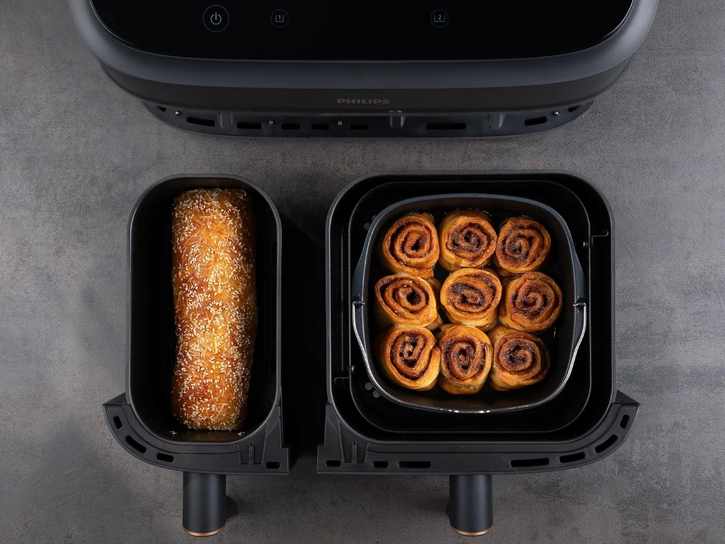 Airfryer Philips Ovi Smart 2.0 dostępny jest w wersji Dual Basket Widok na otwartą frytkownicę beztłuszczową z dwoma pojemnikami, w jednym znajduje się zapiekana bułka z ziarnami, a w drugim kilka drożdżówek cynamonowych ułożonych w okrąg.