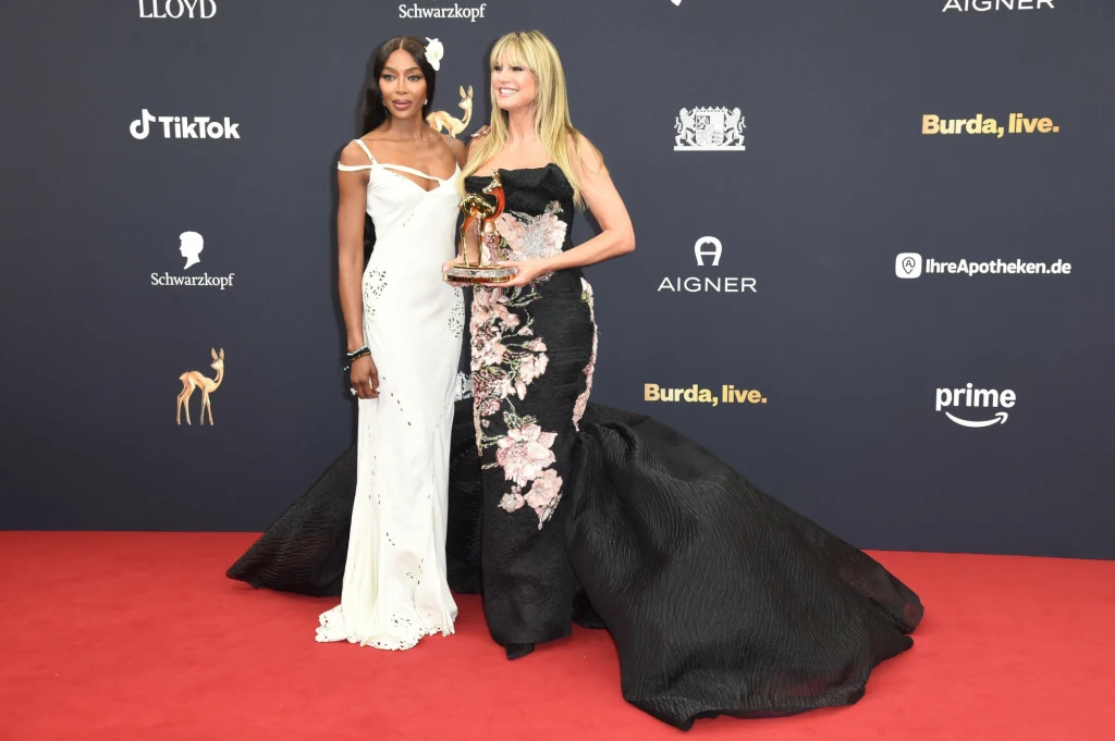 Naomi Campbell i Heidi Klum znów razem Dwie kobiety w eleganckich, wieczorowych sukniach stoją na czerwonym dywanie podczas gali, jedna z nich trzyma statuetkę. W tle widoczne są logotypy sponsorów i partnerów wydarzenia.