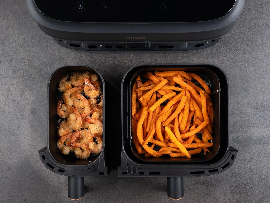 Airfryer Philips Ovi Smart 2.0 dostępny jest w wersji Dual Basket Krewetki panierowane w jednej komorze frytkownicy beztłuszczowej, frytki ze słodkich ziemniaków w drugiej komorze, oba produkty są dokładnie upieczone i apetycznie ułożone na tle czarnej frytkownicy.