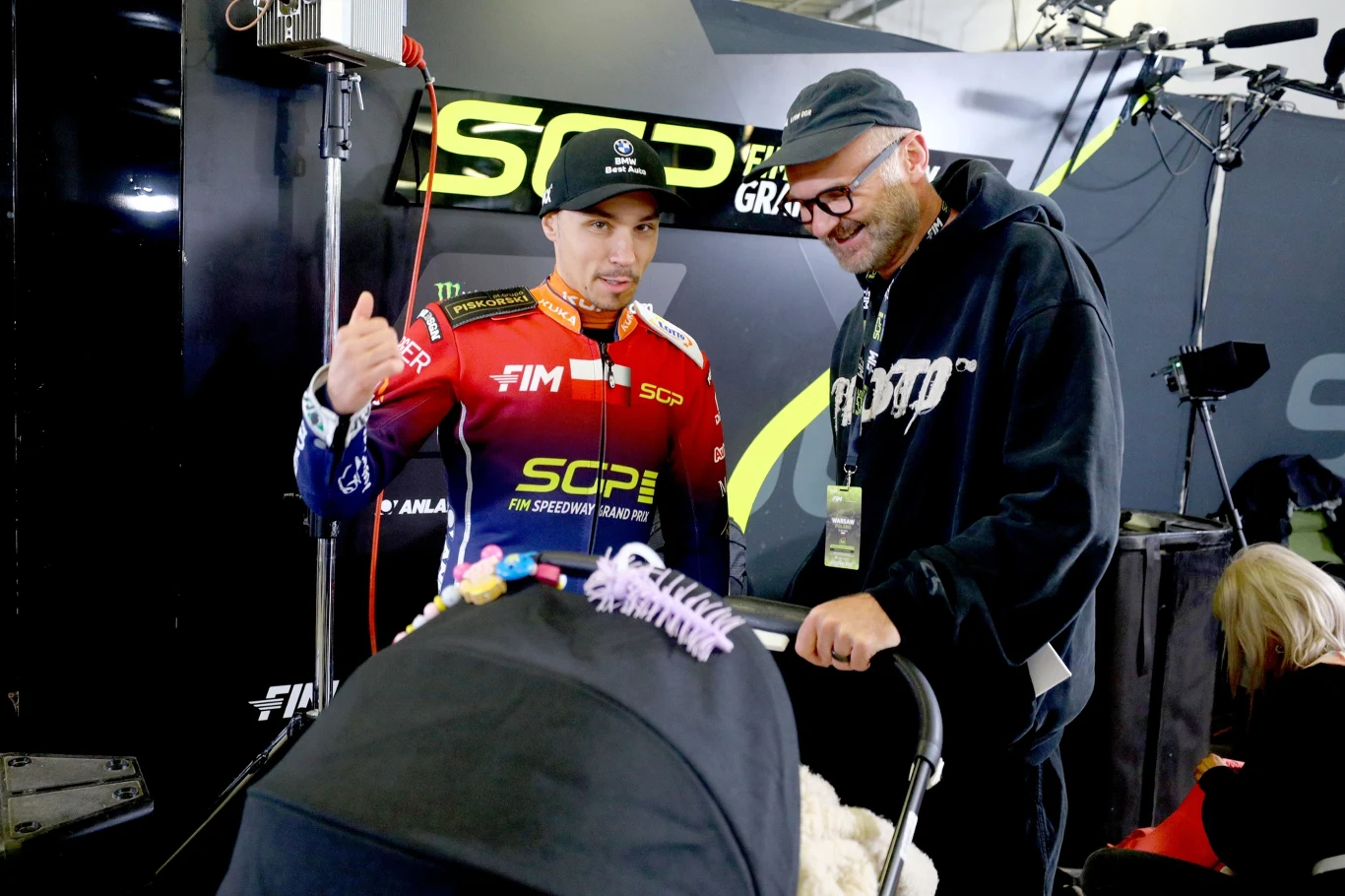 Dwóch mężczyzn dyskutuje w otoczeniu technicznym, jeden ubrany w strój wyścigowy z logotypami sponsora FIM, drugi w ciemnej bluzie z okularami i czapką, na pierwszym planie widoczny wózek dziecięcy ozdobiony kolorowymi zabawkami.