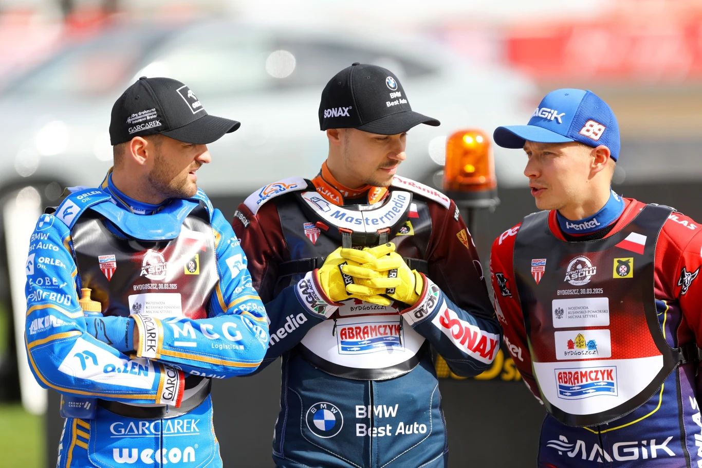 Trzech żużlowców w kolorowych kombinezonach sportowych rozmawia ze sobą na tle toru żużlowego. Każdy zawodnik ubrany jest w specjalistyczny kombinezon z logotypami sponsorów, kaski w rękach lub na głowach, w tle widoczny fragment stadionu.