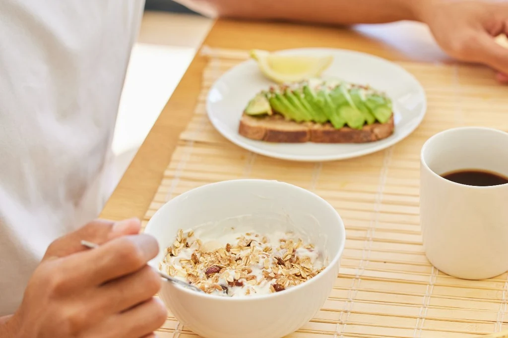 Zadbaj o to, by śniadanie było zbilansowane Śniadanie składające się z miski jogurtu lub mleka z musli, kromki chleba z awokado i kawałkiem cytryny oraz filiżanki czarnej kawy ustawionych na bambusowej podkładce. W kadrze widoczna część osoby sięgającej po jedzenie.