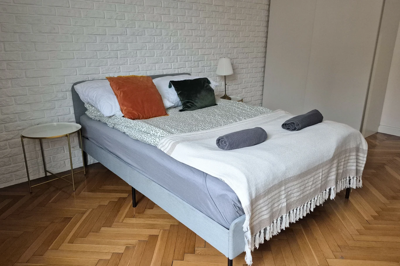 Booking nie nadzoruje procesu uiszczania depozytów i ich zwrotu. Zdjęcie ilustracyjne Łóżko z szarą ramą ustawione przy ścianie z białej cegły, przykryte wzorzystą pościelą i białym kocem, na którym ułożone są dwa zrolowane ręczniki. Na łóżku znajdują się trzy poduszki: biała, zielona i pomarańczowa. Obok łóżka stoi okrągły, złoty stoli...