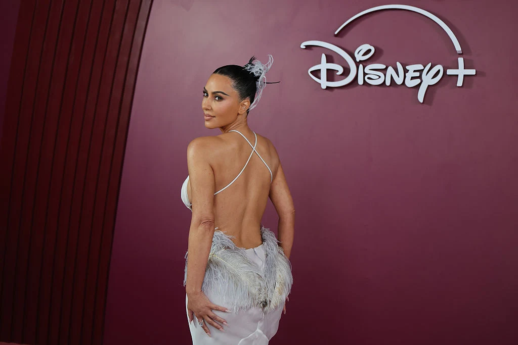 Kim Kardashian Kobieta w eleganckiej, ozdobnej sukni z odkrytymi plecami pozuje na tle bordowej ściany z dużym logo Disney+.