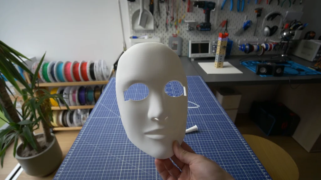 Maskę autor wydrukował na drukarce 3D Biała maska trzymana w dłoni nad niebieską siatką do cięcia, w tle widoczne uporządkowane narzędzia, półki z filamentami do drukarki 3D oraz roślina doniczkowa.