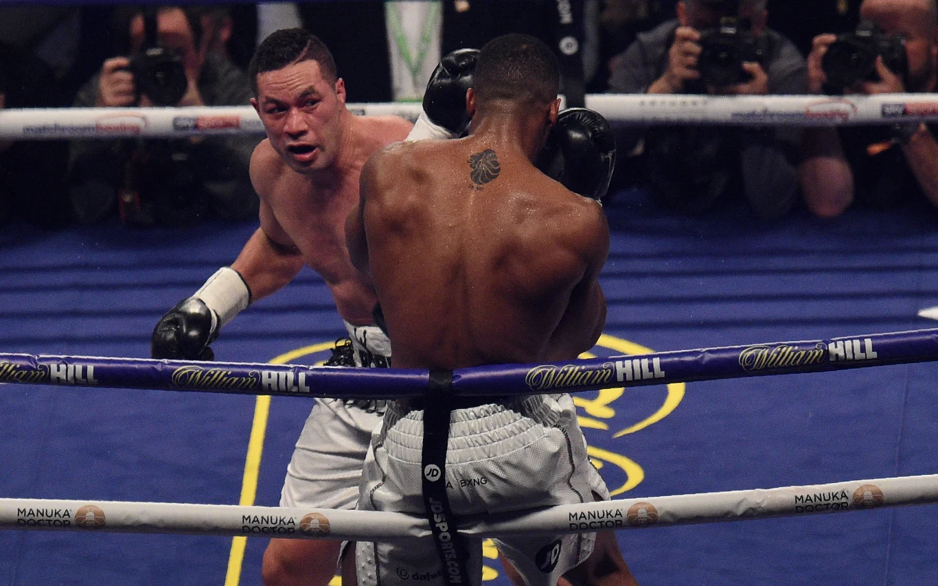 Joseph Parker Dwóch bokserów walczących na ringu, jeden z zawodników atakuje drugim ciosem w kierunku głowy, w tle widownia oraz fotografowie dokumentujący wydarzenie.