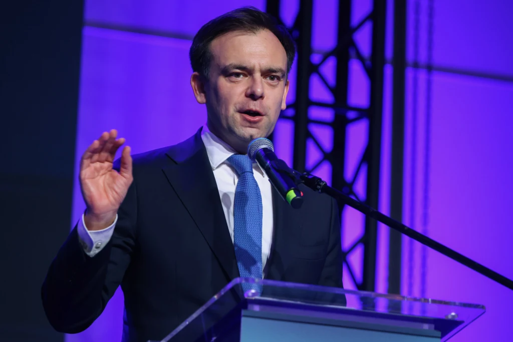 Mężczyzna w garniturze przemawia do mikrofonu stojąc przy przezroczystej mównicy na tle niebiesko-fioletowego oświetlenia.