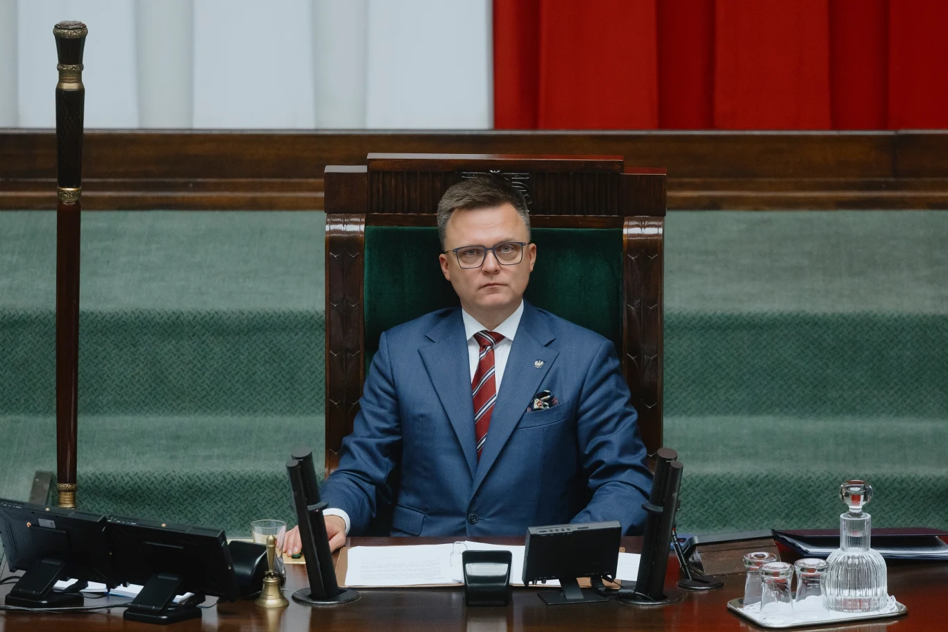 Szymon Hołownia na krótkiej liście kandydatów na komisarza ONZ Mężczyzna w garniturze siedzi za dużym biurkiem w sali obrad, w tle widoczne fragmenty biało-czerwonej flagi, na stole stoją mikrofony, szklanki i dzbanki na wodę.