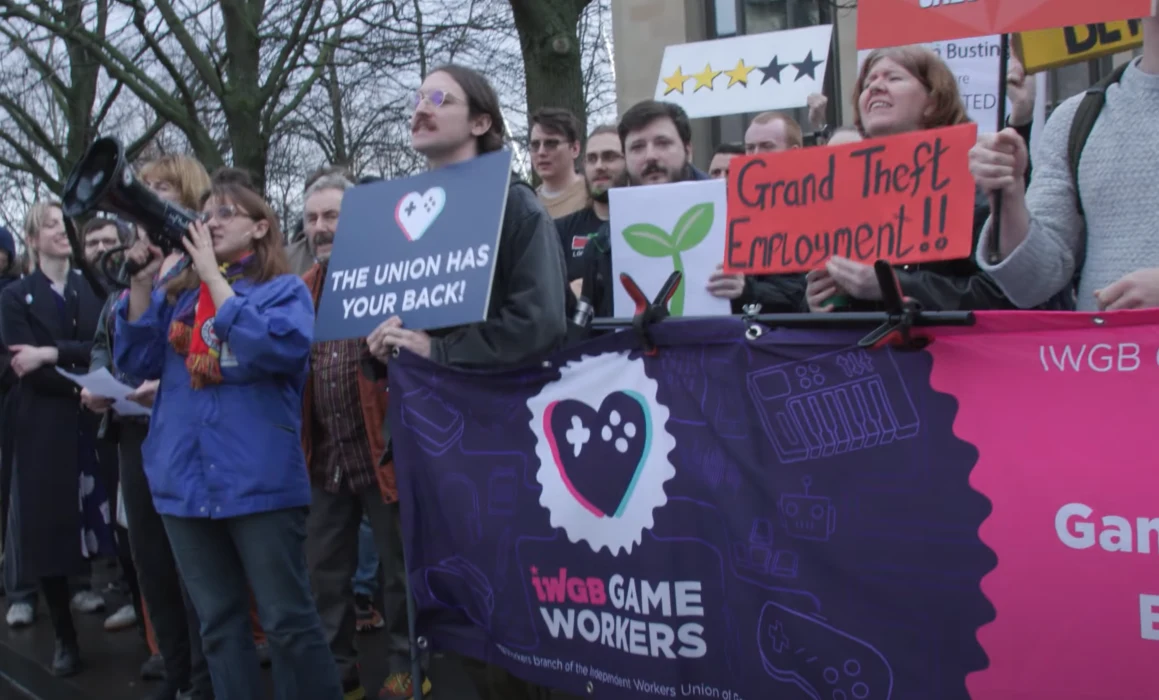 pikieta przed siedzibą Rockstar North / źródło: People Make Games Grupa osób protestujących z transparentami i banerami, część trzyma hasła wspierające związki zawodowe i zatrudnienie w branży gier komputerowych, na pierwszym planie widoczny baner IWGB Game Workers, w tle drzewa i budynki.