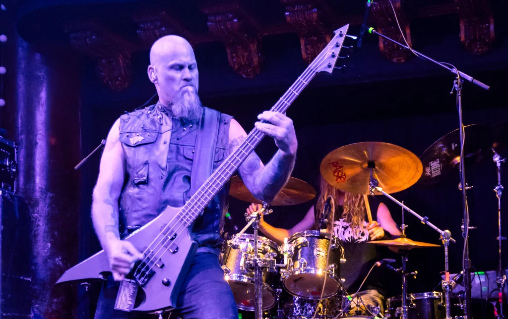 Steve Tucker wszedł w skład supergrupy Malefic Throne Mężczyzna grający na gitarze basowej w klimacie rockowym, u boku perkusista podczas koncertu na scenie oświetlonej kolorowymi światłami.