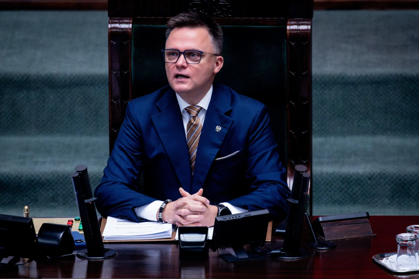 Szymon Hołownia zrezygnował z marszałkowskiego fotela, ale finał jego szefowania Sejmem mógł przebiec inaczej Mężczyzna w garniturze i okularach siedzi przy dużym biurku, prawdopodobnie na stanowisku przewodniczącego w parlamencie. Przed nim znajdują się dokumenty, mikrofony i szklanka wody. Scena ma oficjalny, formalny charakter.
