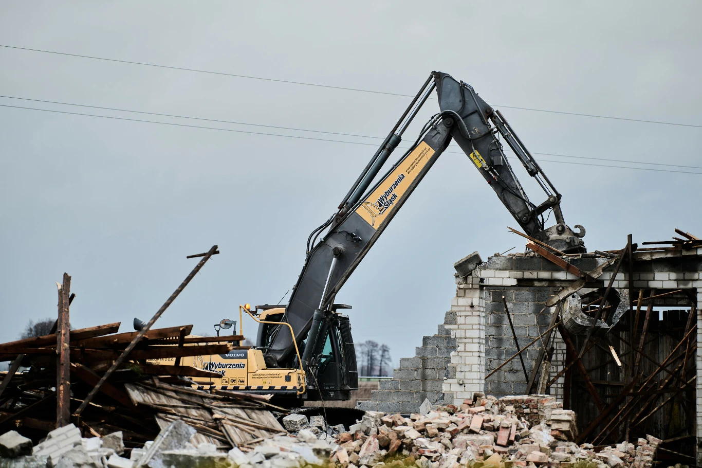 Nowe wywłaszczenia pod CPK Koparka z wysięgnikiem niszczy fragment murowanego budynku, wokół leżą gruz i elementy konstrukcyjne. W tle widoczny jest pochmurny krajobraz, a pracująca maszyna dominuje w centrum ujęcia.