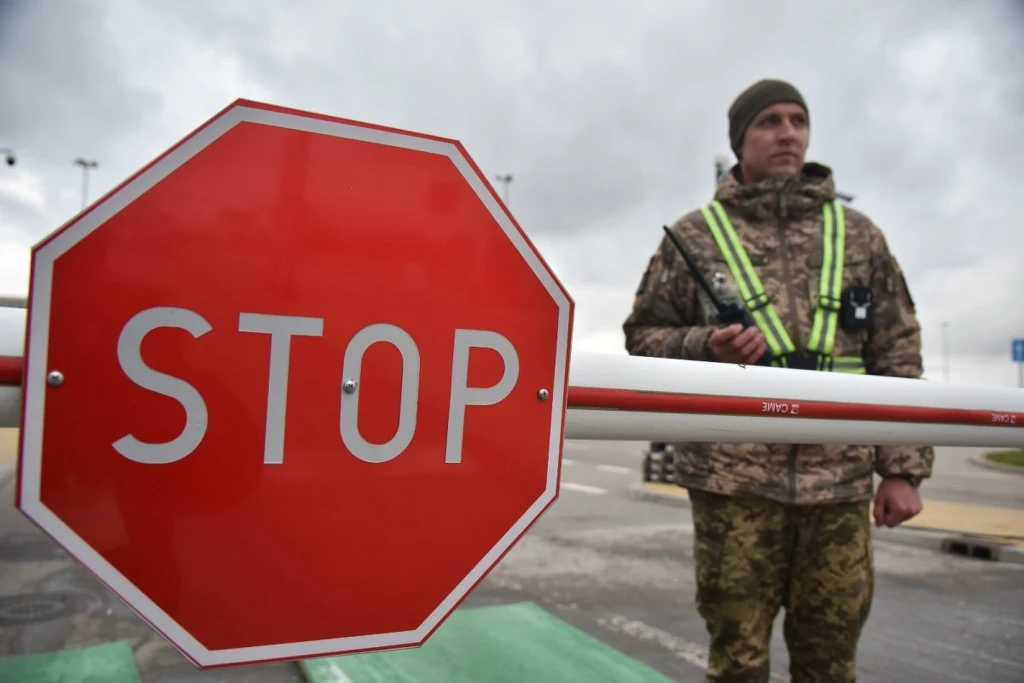 Przejście graniczne z Polską. Zdjęcie ilustracyjne Żołnierz w mundurze stoi przy zamkniętym szlabanie i znaku stop na przejściu granicznym, tło stanowi pochmurne niebo i asfaltowa nawierzchnia.