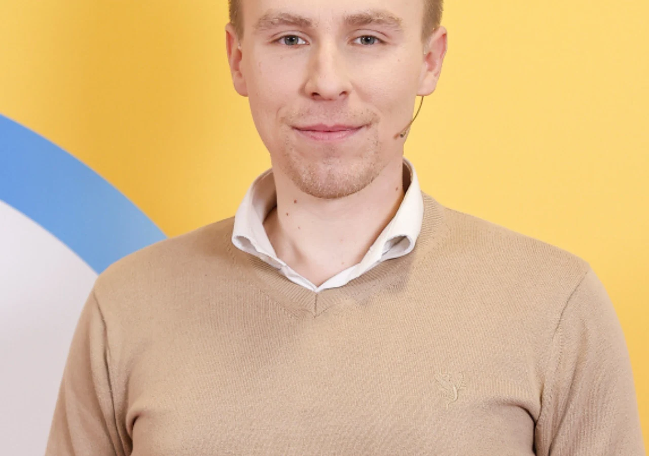 Bartosz Radziejewski - zwycięzca "Milionerów" Młody mężczyzna z mikrofonem przy twarzy, ubrany w jasnobrązowy sweter i białą koszulę, stoi na tle żółtej ściany z fragmentem niebiesko-białego elementu po lewej stronie kadru.