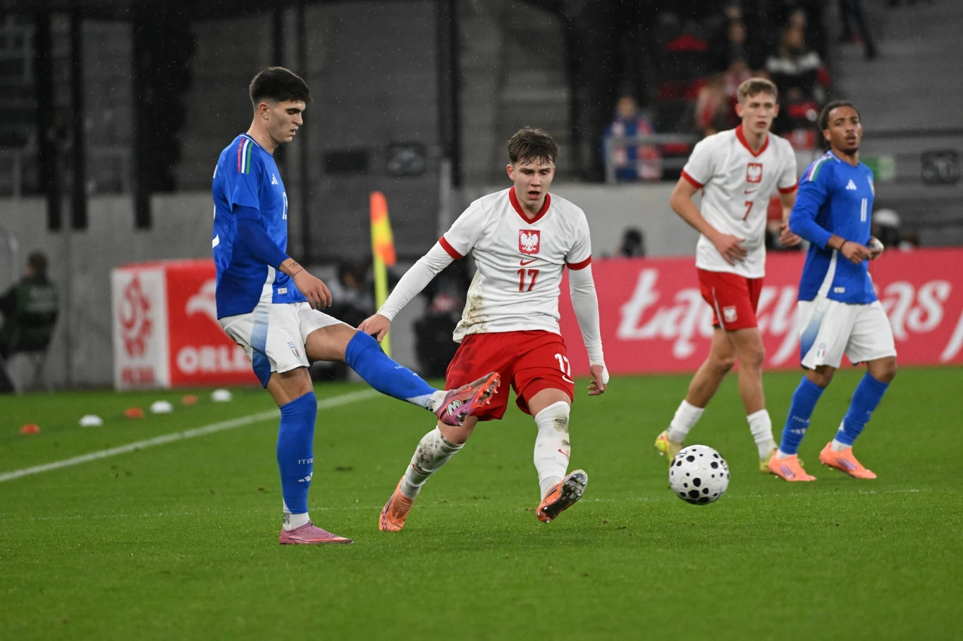 Polska U21 - Włochy U21 Piłkarz w niebieskim stroju próbuje odebrać piłkę zawodnikowi z numerem 17 w białych barwach z czerwonymi elementami. W tle inni zawodnicy oraz fragment trybun stadionu.