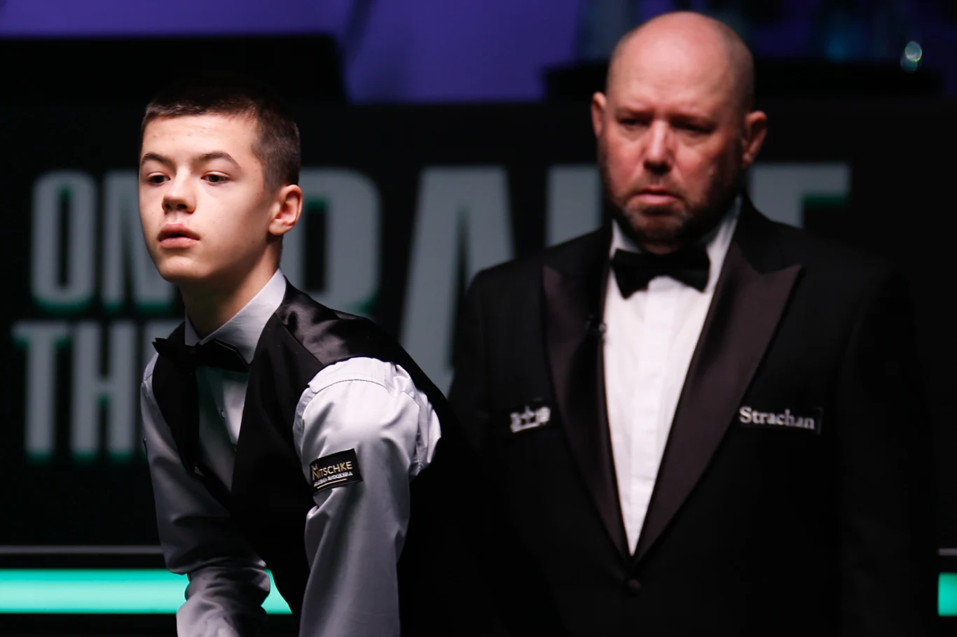 14-letni Michał Szubarczyk już za kilka dni zmierzy się z legendą snookera Młody snookerzysta w skupieniu przygotowuje się do uderzenia, obok stoi sędzia w czarnym garniturze i muszce, obaj ubrani w oficjalne stroje turniejowe, na ciemnym tle z elementami scenografii turnieju.