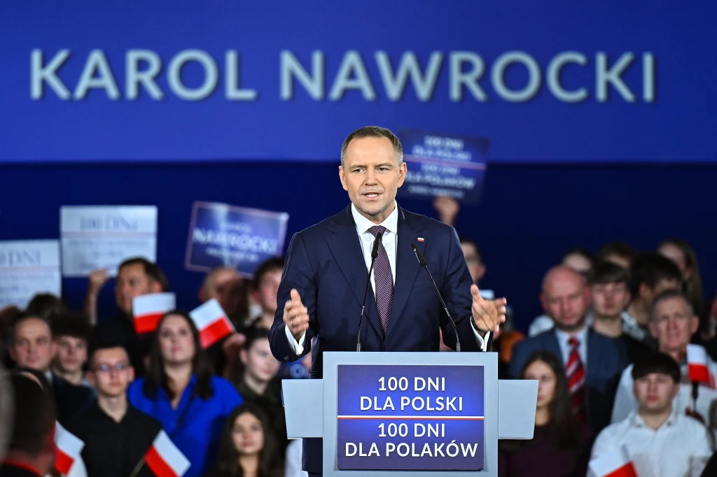 Prezydent Karol Nawrocki Mężczyzna w garniturze przemawia na tle niebieskiego baneru z napisem 'Karol Nawrocki'. Za nim tłum osób trzymających polskie flagi i plakaty z napisem '100 dni dla Polski 100 dni dla Polaków'.