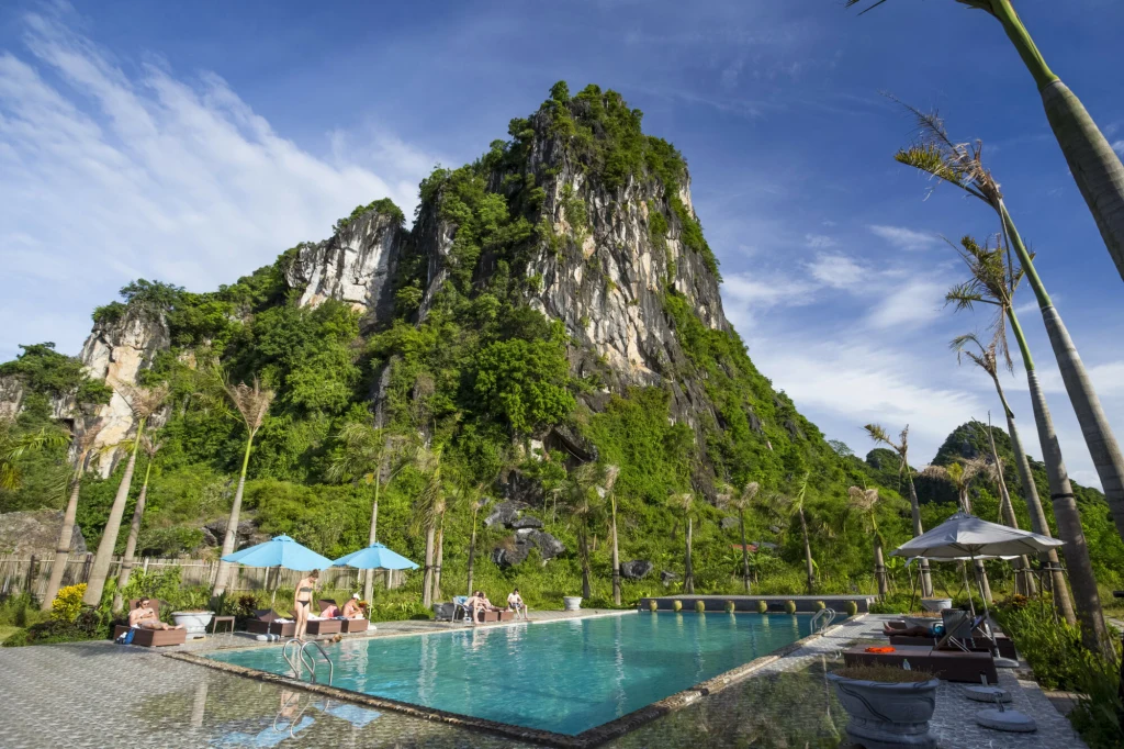 Widok na park narodowy Phong Nha-Kẻ Bàng Basen otoczony palmami, z ludźmi odpoczywającymi na leżakach, w tle imponująca, zielona skała górska oraz błękitne niebo z delikatnymi chmurami.