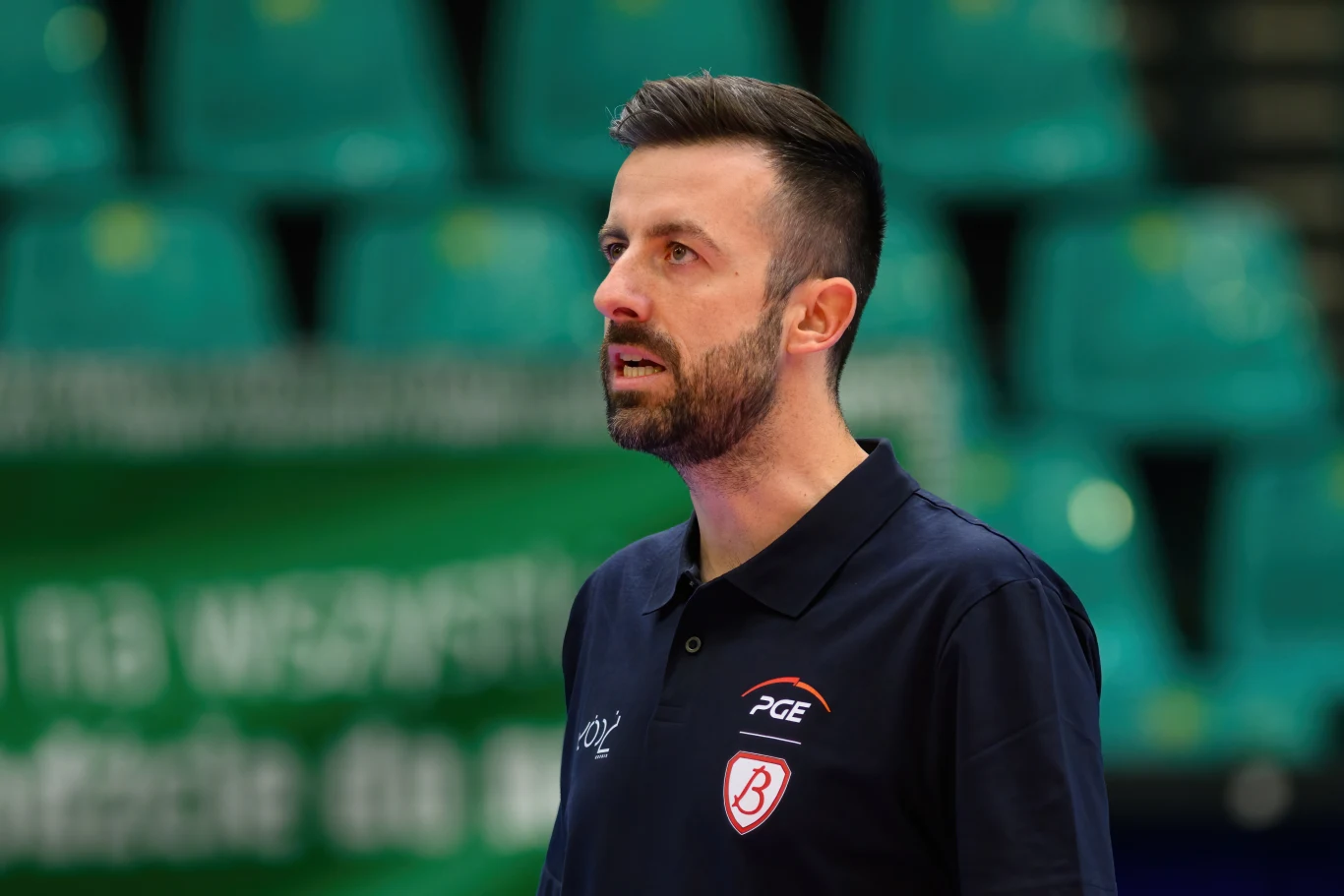 Trener w granatowej koszulce z logo klubu sportowego na tle pustych zielonych trybun hali sportowej.