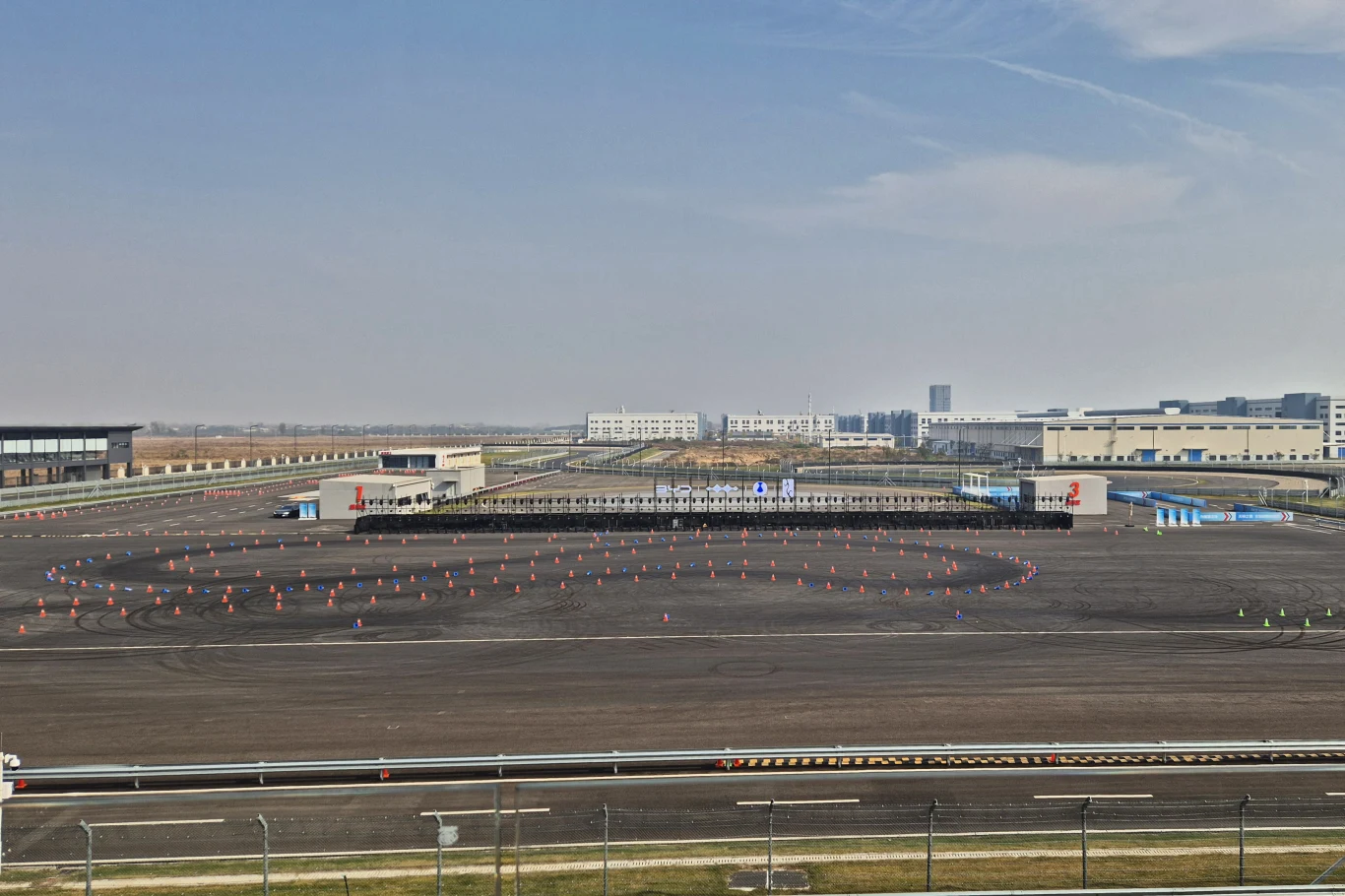 Ośrodek testowy BYD w Zhengzhou Duży otwarty plac asfaltowy z rozmieszczonymi pachołkami tworzącymi owalny tor testowy, w tle nowoczesne budynki przemysłowe i infrastruktura techniczna. Przed polem widoczny płot zabezpieczający miejsce.