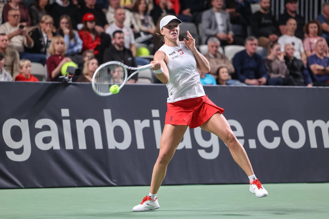 Tenisistka w białej koszulce i czerwonej spódniczce wykonuje uderzenie podczas meczu na korcie twardym, w tle widownia uważnie obserwuje rozgrywkę.