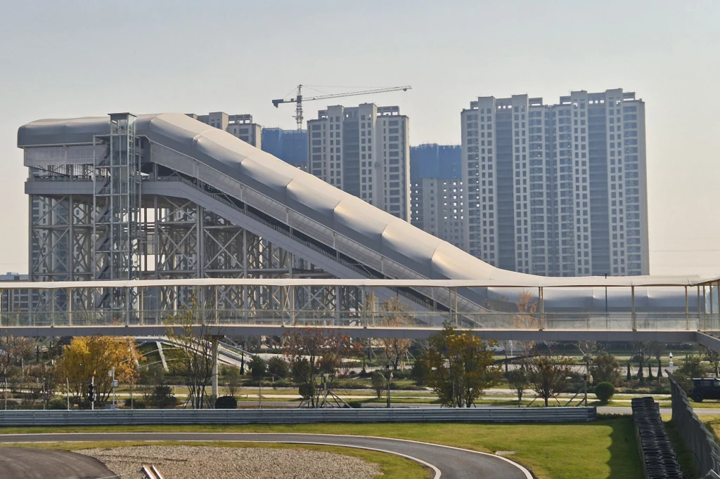 Ta nietypowa konstrukcja kryje największą na świecie sztuczną wydmę do testów samochodów Nowoczesna skocznia narciarska o stalowej konstrukcji w otoczeniu zieleni, w tle wysokie budynki mieszkalne oraz dźwig budowlany. Przestrzeń podzielona na kilka poziomych warstw, a urbanistyczny krajobraz podkreślony przez obecność roślinności i dróg.